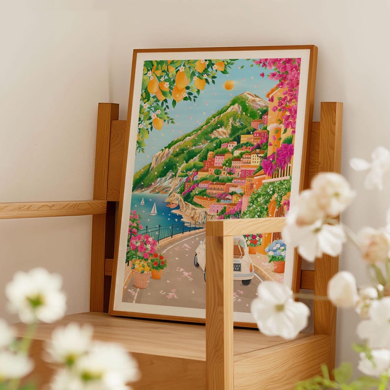 May include: A framed art print depicting a coastal Italian town. The artwork features colorful buildings on a hillside, a road with a vintage car, and a blue sea with sailboats. Lemon trees and pink flowers frame the scene.