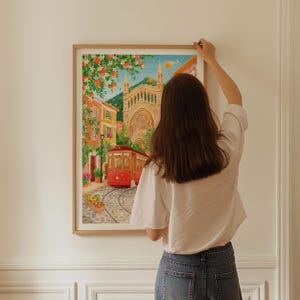 May include: A framed watercolor print depicting a European city scene with a red trolley car on cobblestone tracks. The artwork features colorful buildings, lush greenery, and a large cathedral in the background. The print is in a light wood frame.