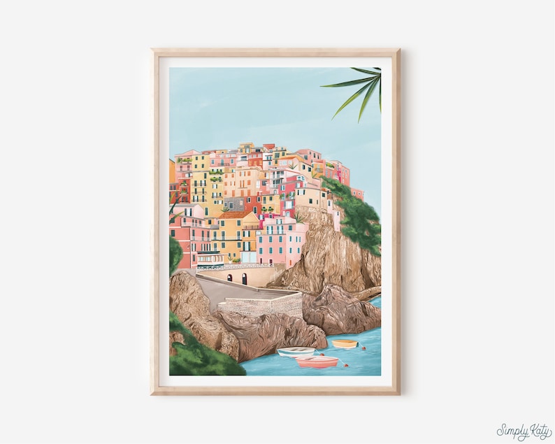May include: A framed art print depicts a colorful coastal village scene. The buildings are painted in shades of pink, yellow, and orange, set against a light blue sky and turquoise water. Small boats float in the foreground. The print is in a light wood frame.
