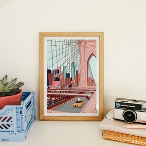 May include: A framed art print depicts a stylized illustration of the Brooklyn Bridge with a yellow taxi cab. The artwork features a color palette of pink, blue, and brown tones. A succulent plant in a pink pot and a vintage camera are also visible.