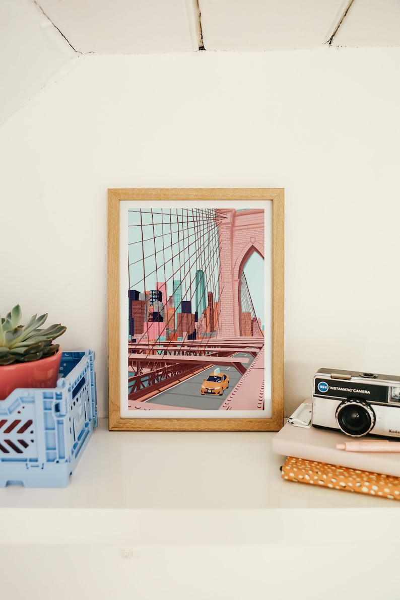 May include: A framed art print depicts a stylized illustration of the Brooklyn Bridge with a yellow taxi cab. The artwork features a color palette of pink, blue, and brown tones. A succulent plant in a pink pot and a vintage camera are also visible.