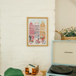 Puede incluir: Una impresi&oacute;n enmarcada de una ilustraci&oacute;n colorida de edificios y una bicicleta delante de ellos. La impresi&oacute;n est&aacute; colgada en una pared de ladrillo blanco. Un tocadiscos vintage est&aacute; sobre una mesa de madera delante de la impresi&oacute;n.