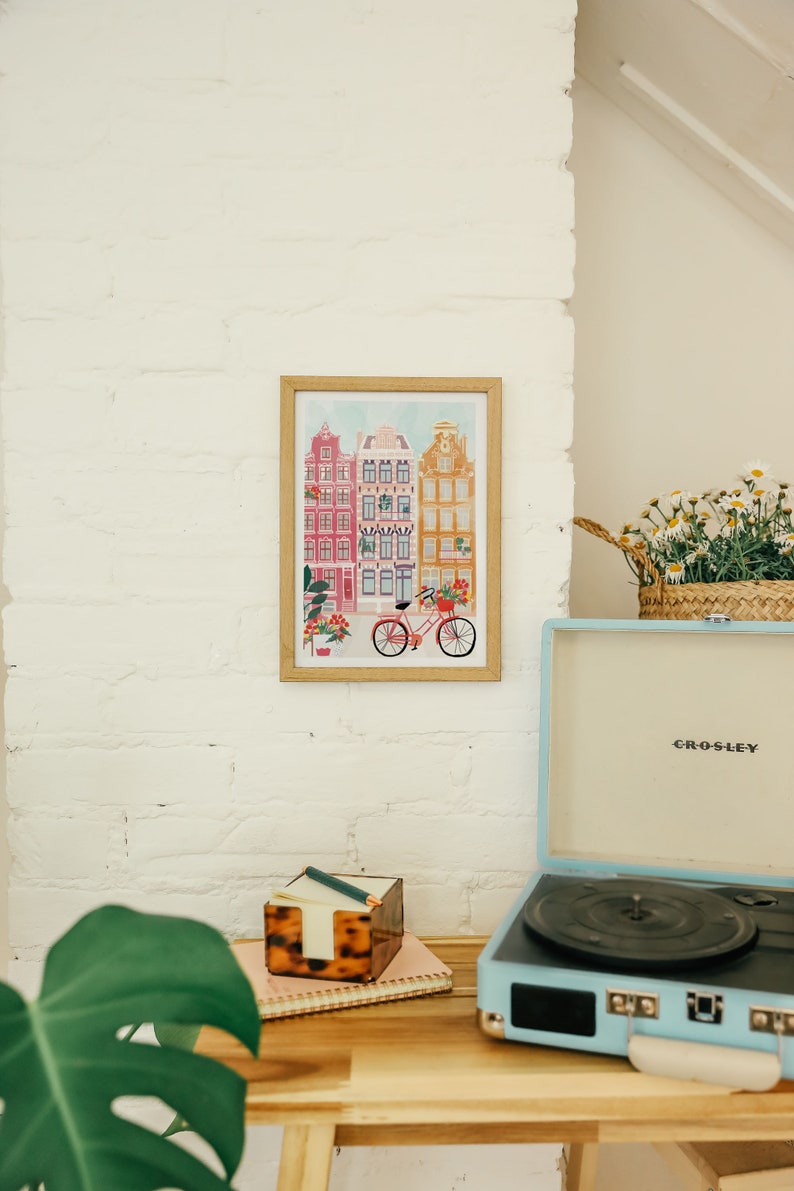 Puede incluir: Una impresi&oacute;n enmarcada de una ilustraci&oacute;n colorida de edificios y una bicicleta delante de ellos. La impresi&oacute;n est&aacute; colgada en una pared de ladrillo blanco. Un tocadiscos vintage est&aacute; sobre una mesa de madera delante de la impresi&oacute;n.