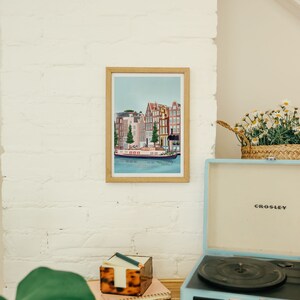May include: A framed illustration of a canal scene in Amsterdam, Netherlands. The illustration features colorful buildings, a canal, and a boat. The frame is made of wood and has a light brown finish. The illustration is hanging on a white brick wall. A vintage record player is on a wooden table in the foreground.