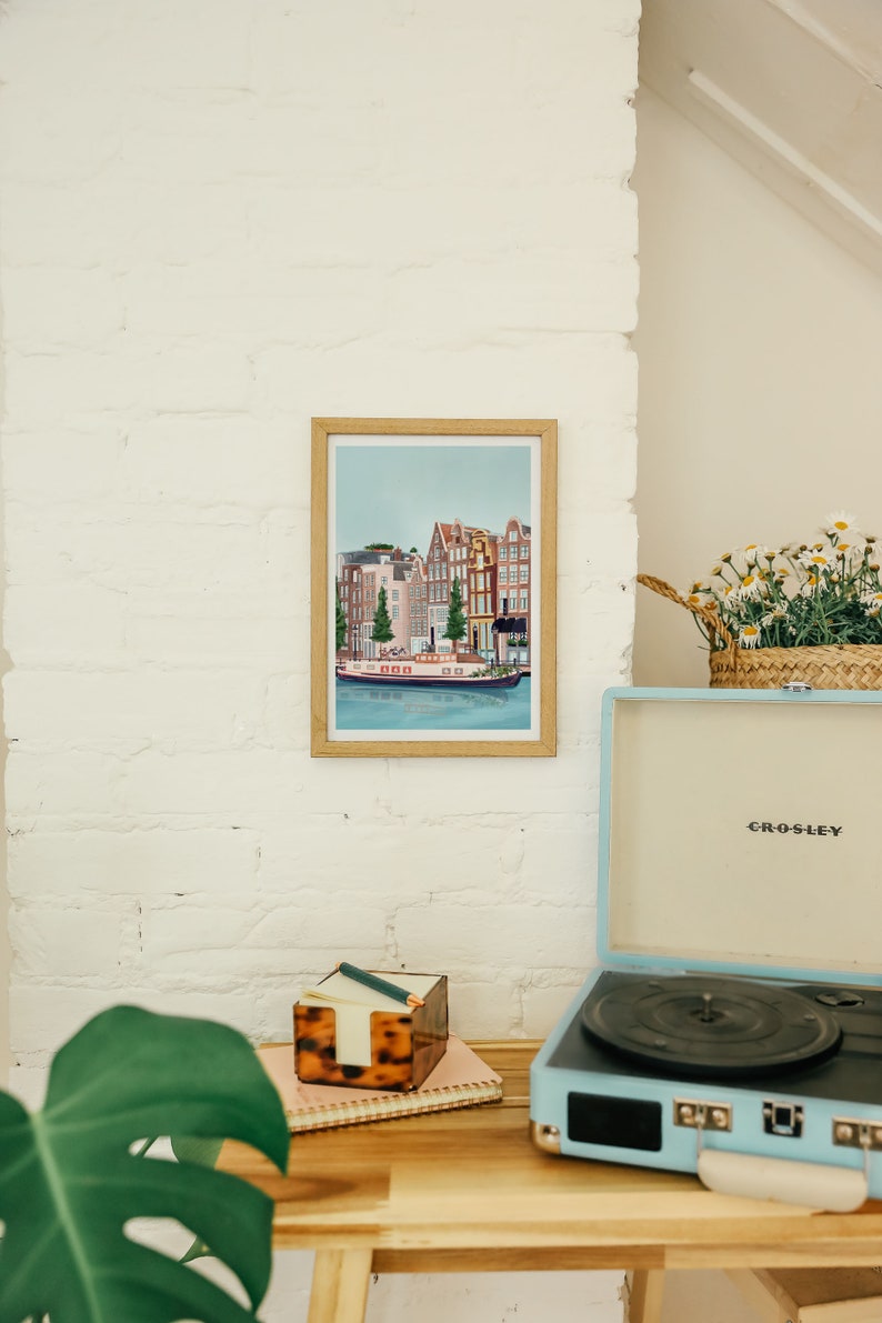 May include: A framed illustration of a canal scene in Amsterdam, Netherlands. The illustration features colorful buildings, a canal, and a boat. The frame is made of wood and has a light brown finish. The illustration is hanging on a white brick wall. A vintage record player is on a wooden table in the foreground.