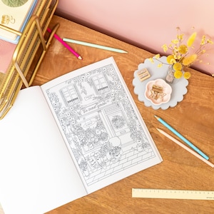 May include: An open coloring book featuring a detailed black and white illustration of a flower shop with the text "Le Petit Jardin". Colored pencils, a gold desk organizer, and decorative items are arranged on a wooden surface.