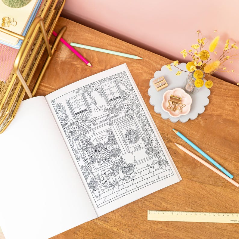 May include: An open coloring book featuring a detailed black and white illustration of a flower shop with the text "Le Petit Jardin". Colored pencils, a gold desk organizer, and decorative items are arranged on a wooden surface.