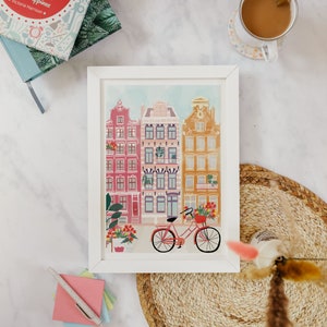 Puede incluir: Impresi&oacute;n enmarcada en blanco de una ilustraci&oacute;n colorida de una escena de calle con edificios y una bicicleta. Los edificios son de color rosa, rojo y amarillo con molduras blancas. La bicicleta es roja con una cesta de flores.