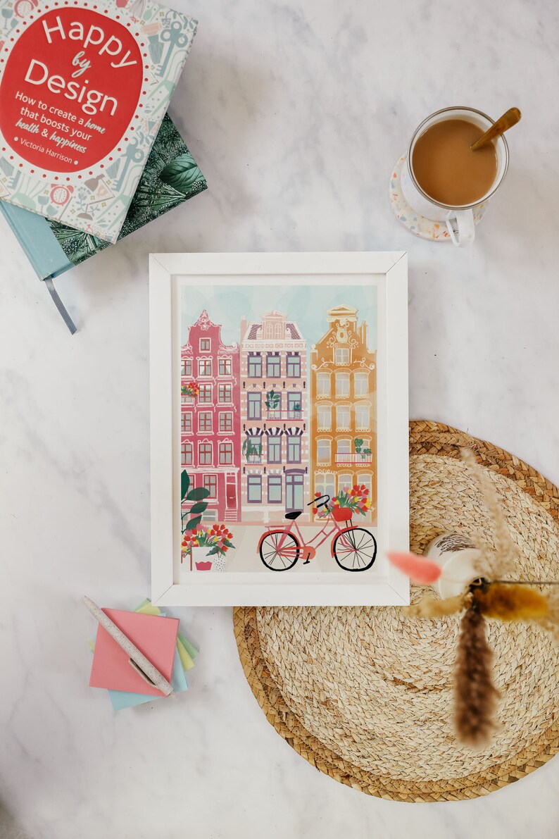 Puede incluir: Impresi&oacute;n enmarcada en blanco de una ilustraci&oacute;n colorida de una escena de calle con edificios y una bicicleta. Los edificios son de color rosa, rojo y amarillo con molduras blancas. La bicicleta es roja con una cesta de flores.