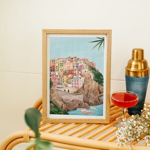 May include: A framed art print depicts a colorful coastal village nestled on a cliffside, with boats in the water. A cocktail and shaker sit nearby on a rattan table, with sprigs of white flowers.