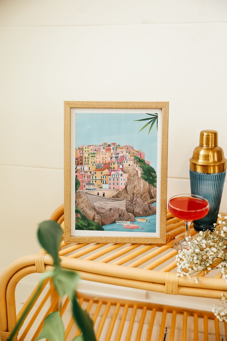 May include: A framed art print depicts a colorful coastal village nestled on a cliffside, with boats in the water. A cocktail and shaker sit nearby on a rattan table, with sprigs of white flowers.