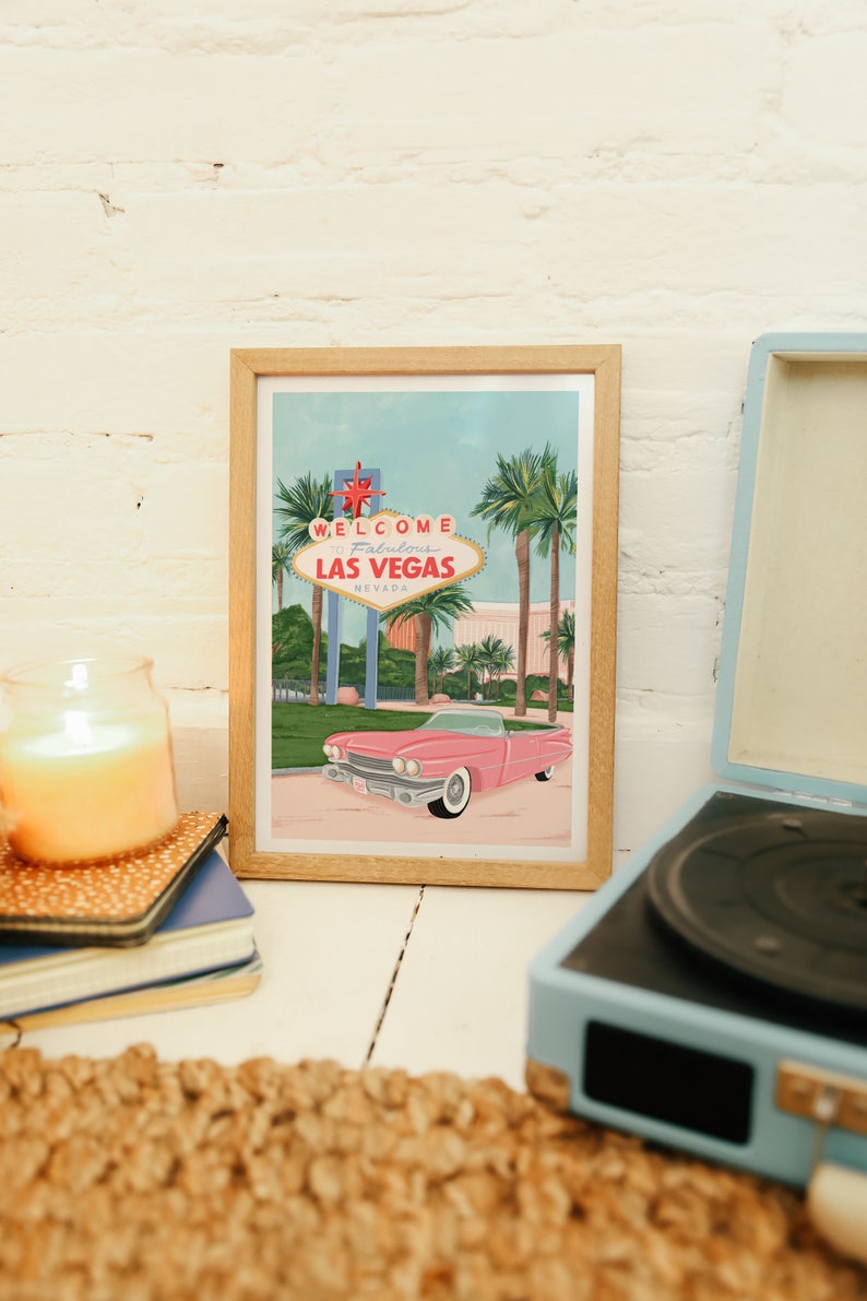 May include: A framed illustration of the "Welcome to Fabulous Las Vegas" sign with a pink vintage car in front of it. The illustration is set against a blue sky with palm trees.
