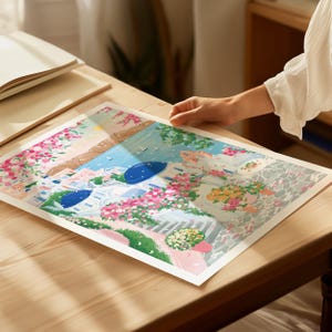 以下が含まれることがあります： 白い建物、青いドーム、鮮やかなピンクと黄色の花が描かれた、カラフルな沿岸の町のプリント。 このアートワークは、青い空と海のある晴れた日を描いており、陽気なシーンを作り出しています。 プリントは木製のテーブルに置かれています。
