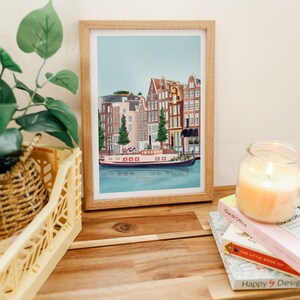 May include: A framed illustration of a canal in Amsterdam, Netherlands. The illustration features colorful buildings with red tile roofs and a boat on the water. The illustration is in a light wood frame.