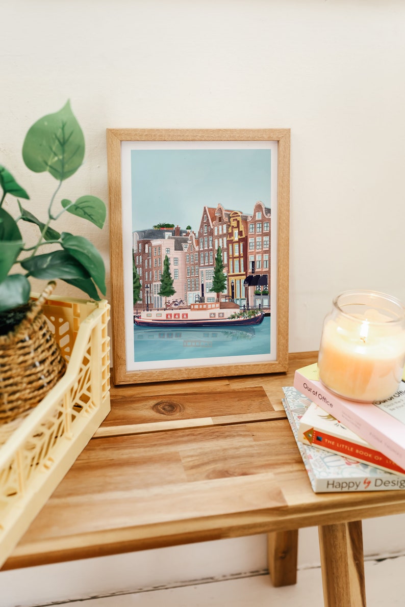 May include: A framed illustration of a canal in Amsterdam, Netherlands. The illustration features colorful buildings with red tile roofs and a boat on the water. The illustration is in a light wood frame.