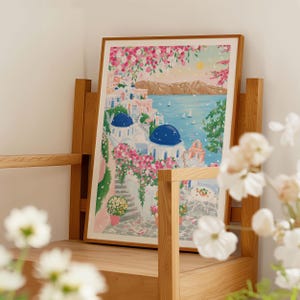 以下が含まれることがあります： 白い建物、青いドーム、ピンクの花が描かれた、海岸沿いの村の風景を描いた額入りのアートプリント。青い海と黄色の太陽を背景に、木製の棚に飾られています。
