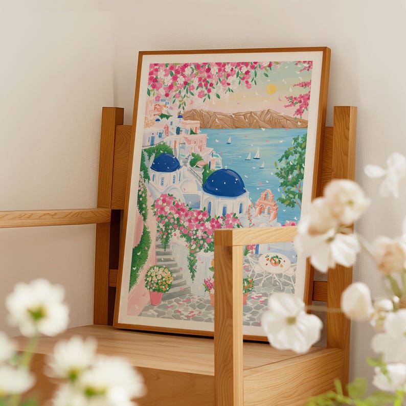 以下が含まれることがあります： 白い建物、青いドーム、ピンクの花が描かれた、海岸沿いの村の風景を描いた額入りのアートプリント。青い海と黄色の太陽を背景に、木製の棚に飾られています。