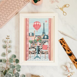 May include: A framed illustration of a Parisian balcony with a hot air balloon, Eiffel Tower, and a table with two chairs. The balcony is decorated with pink and white flowers. The illustration is in a white frame.