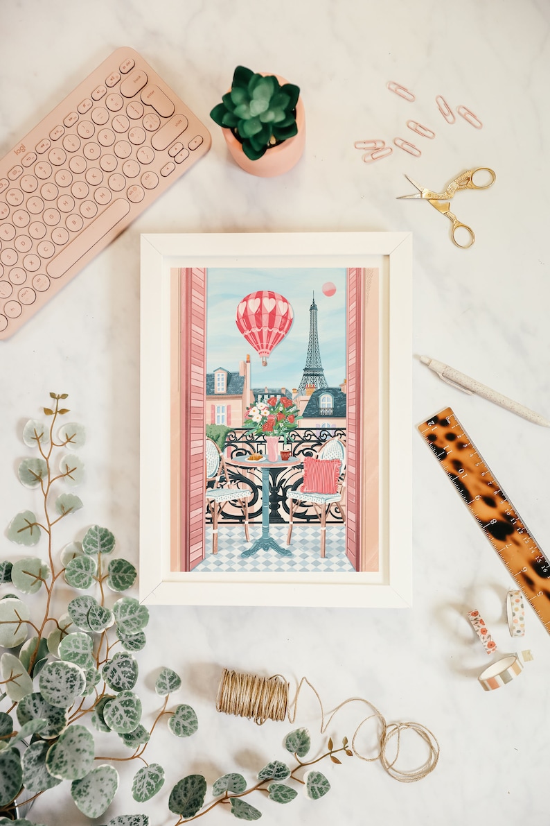 May include: A framed illustration of a Parisian balcony with a hot air balloon, Eiffel Tower, and a table with two chairs. The balcony is decorated with pink and white flowers. The illustration is in a white frame.