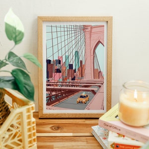 May include: A framed art print depicting the Brooklyn Bridge in New York City. The illustration features a yellow taxi cab driving across the bridge, with the city skyline in the background. The print is in a wooden frame and is displayed on a wooden surface.