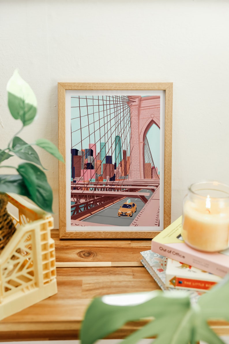 May include: A framed art print depicting the Brooklyn Bridge in New York City. The illustration features a yellow taxi cab driving across the bridge, with the city skyline in the background. The print is in a wooden frame and is displayed on a wooden surface.