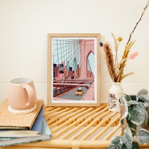 May include: A framed art print depicting the Brooklyn Bridge with a yellow taxi cab driving across. The artwork features a pink and blue color palette, with a light wood frame. The print is displayed on a woven shelf with a pink mug and books.