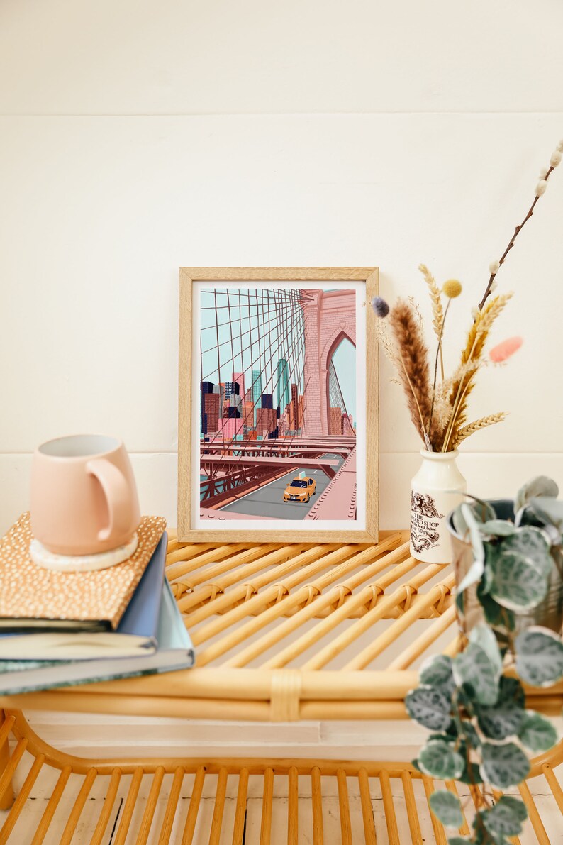 May include: A framed art print depicting the Brooklyn Bridge with a yellow taxi cab driving across. The artwork features a pink and blue color palette, with a light wood frame. The print is displayed on a woven shelf with a pink mug and books.