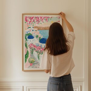 以下が含まれることがあります： 白い建物、青いドーム、ピンクの花が描かれた、海岸の風景の額入りアートプリント。 アートワークは明るい木製のフレームに入っています。 画像は、白い壁に額入りのアートを掛けている人を示しています。