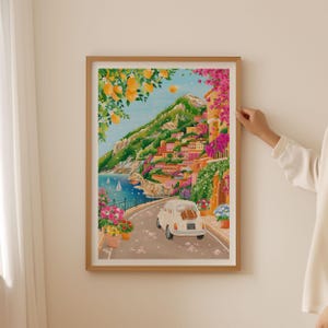 May include: A framed art print depicting a coastal Italian town. The artwork features a winding road, a white car, colorful buildings, and lush greenery. The scene is framed by a wooden border, creating a vibrant and inviting aesthetic.