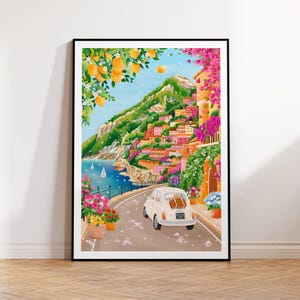 May include: A framed art print depicting a coastal Italian town. The artwork features a white car driving on a road with colorful buildings, lush greenery, and a view of the sea. Lemon trees and pink flowers frame the scene.