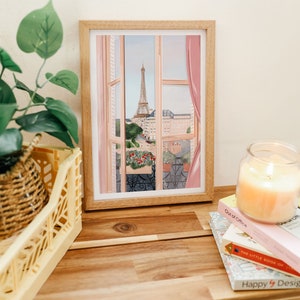 Op de afbeelding: Een ingelijste illustratie van een raam met uitzicht op de Eiffeltoren in de verte. Het raam is open en heeft roze gordijnen. De illustratie staat op een houten tafel met boeken en een kaars.