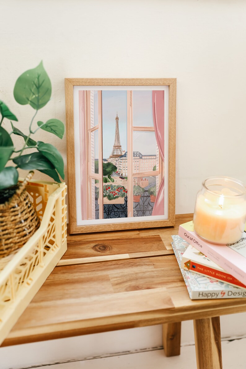 Op de afbeelding: Een ingelijste illustratie van een raam met uitzicht op de Eiffeltoren in de verte. Het raam is open en heeft roze gordijnen. De illustratie staat op een houten tafel met boeken en een kaars.