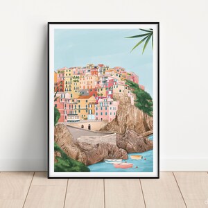 May include: A framed art print depicting a colorful coastal village scene. The artwork showcases buildings in shades of pink, yellow, and orange, set against a light blue sky and turquoise water. Several small boats are visible in the water.