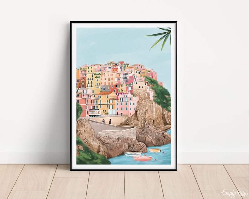 May include: A framed art print depicting a colorful coastal village scene. The artwork showcases buildings in shades of pink, yellow, and orange, set against a light blue sky and turquoise water. Several small boats are visible in the water.