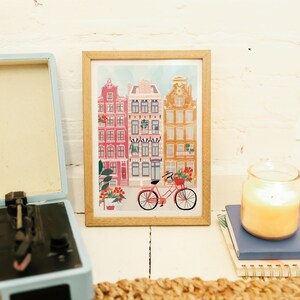 Puede incluir: Una impresi&oacute;n enmarcada de una ilustraci&oacute;n colorida de una fila de edificios con una bicicleta roja en frente. Los edificios est&aacute;n pintados en tonos de rosa, naranja y amarillo. La bicicleta tiene una cesta llena de flores.