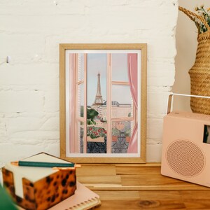 Op de afbeelding: Een ingelijste illustratie van een raam met uitzicht op de Eiffeltoren in Parijs. Het raam is open en de gordijnen zijn opengeschoven. De Eiffeltoren is in de verte en op de voorgrond zijn gebouwen en bomen te zien. De illustratie is in een lichtroze en blauwe kleurstelling.