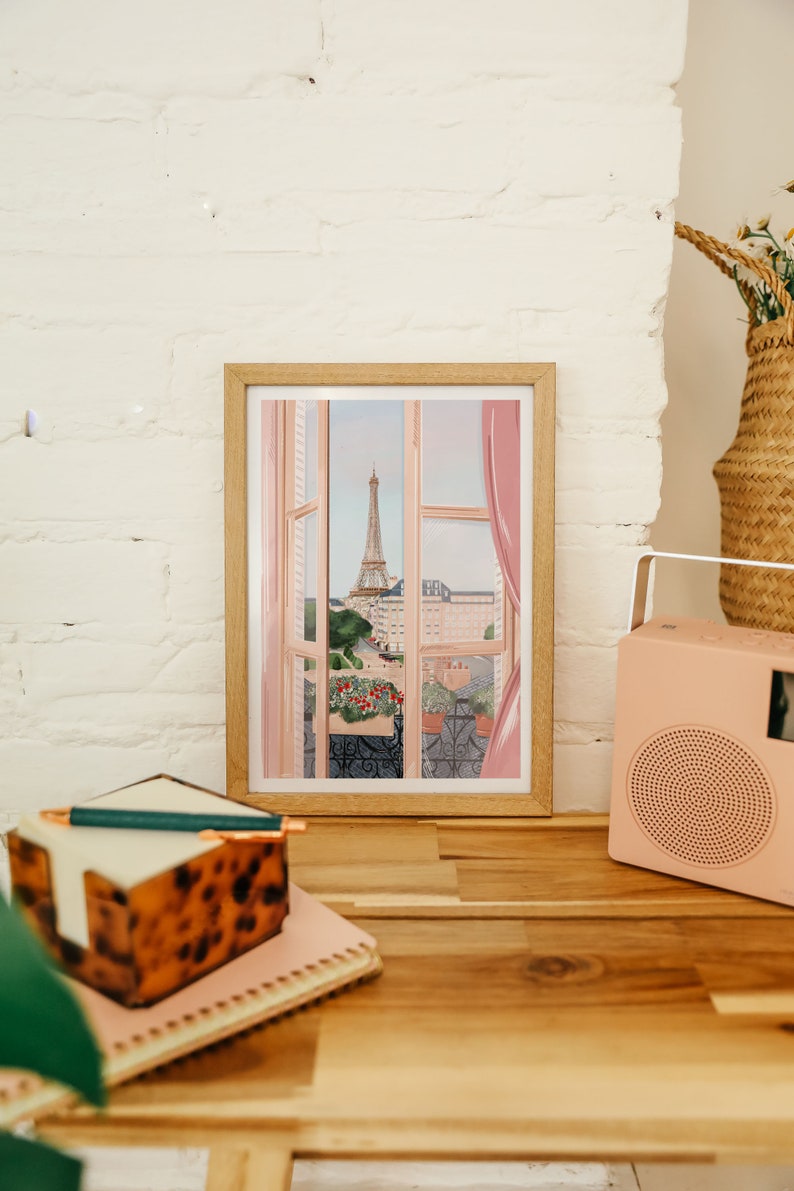 Op de afbeelding: Een ingelijste illustratie van een raam met uitzicht op de Eiffeltoren in Parijs. Het raam is open en de gordijnen zijn opengeschoven. De Eiffeltoren is in de verte en op de voorgrond zijn gebouwen en bomen te zien. De illustratie is in een lichtroze en blauwe kleurstelling.