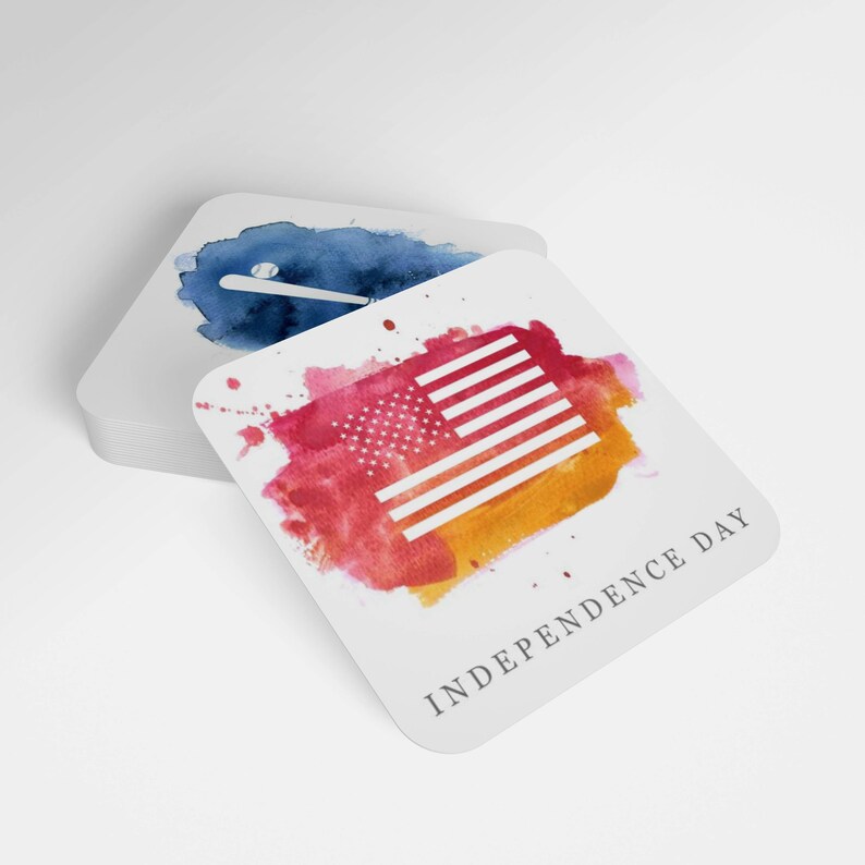 May include: A white square coaster with a red, orange, and yellow watercolor splatter design. The design features a white silhouette of the American flag. The text "INDEPENDENCE DAY" is printed in gray at the bottom of the coaster.