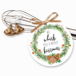 May include: Round white gift tag with a green watercolor wreath, pine cones, and red berries. The text reads "We whisk you a merry kissmas".