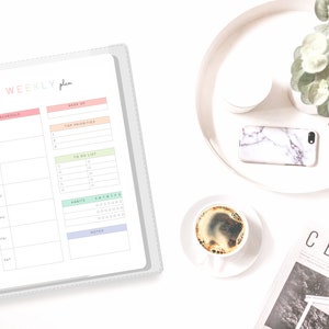 May include: A weekly planner with a gray cover and a white interior. The planner has a schedule section, a top priorities section, a to-do list section, a habits section, and a notes section. The planner is open to a page with the schedule section and the top priorities section. The planner is on a white surface with a cup of coffee, a phone, and a magazine.