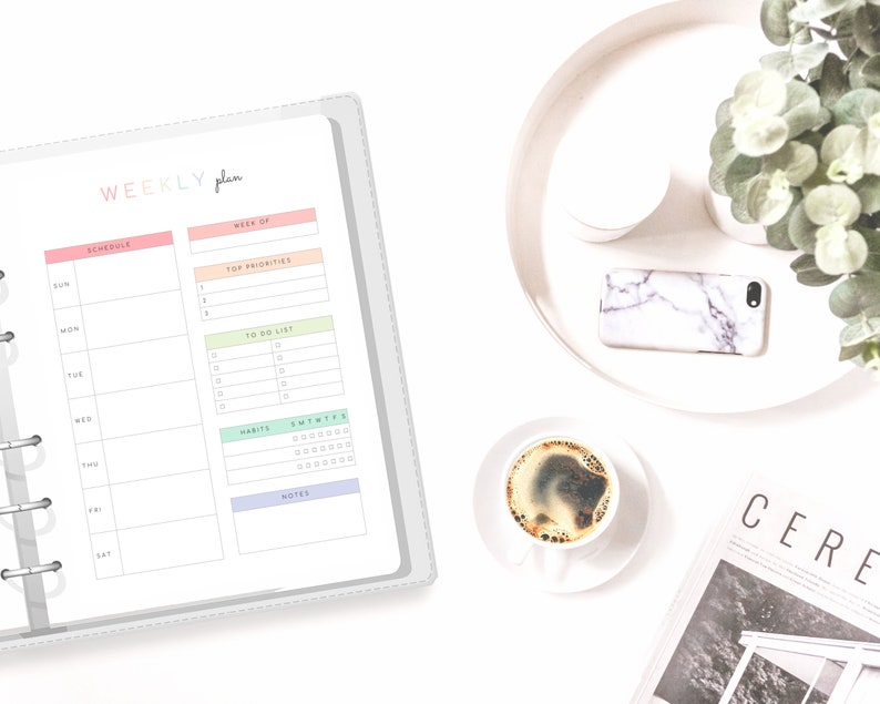 May include: A weekly planner with a gray cover and a white interior. The planner has a schedule section, a top priorities section, a to-do list section, a habits section, and a notes section. The planner is open to a page with the schedule section and the top priorities section. The planner is on a white surface with a cup of coffee, a phone, and a magazine.