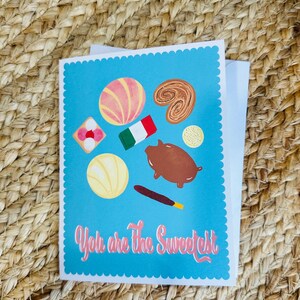 May include: A blue card with a white border and scalloped edges. The card features a variety of colourful Mexican pastries and the text "You are the Sweetest".