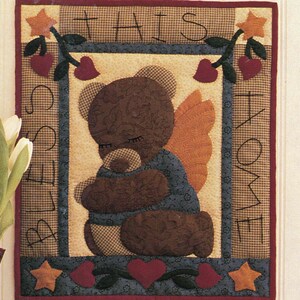 May include: A wallhanging quilt pattern titled Bear Blessing with a finished size of 13 inches by 15 inches. The design features a brown bear with wings, surrounded by a border with the text THIS BLESS HOME, red hearts, and orange stars.