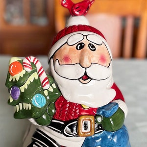 May include: A ceramic Santa Claus figurine, holding a green Christmas tree and a candy cane. The Santa is wearing a red suit with a black belt and a blue sack. A red bird sits atop his hat. The figurine is on a light blue tablecloth.