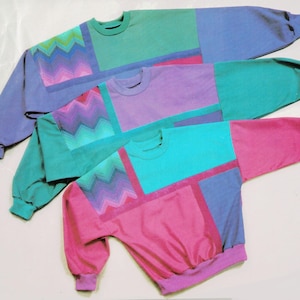 May include: Three colorful sweatshirts in shades of teal, purple, and pink. Each sweatshirt features a color-blocked design with a geometric pattern on the front. The sleeves and body are different colors, creating a vibrant, retro look.