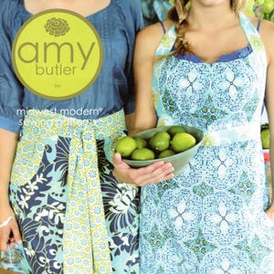 May include: A sewing pattern cover featuring two aprons. The left apron has a blue and green floral pattern with a long tie. The right apron features a light blue and white geometric tile print. Text includes Amy Butler, Midwest Modern, and Domestic Goddess.