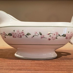 May include: White ceramic gravy boat with a green and pink floral design. The boat has a handle and a wide base.