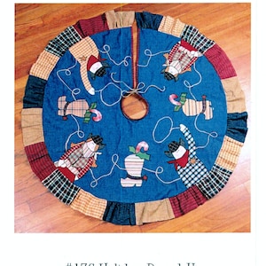 May include: A blue fabric tree skirt featuring appliquéd figures of people in hats and boots, connected by white rope designs. The skirt has a ruffled border made of alternating plaid and checkered fabric patches. Text reads Down Calico Alley #176 Holiday Round-Up Fusible Appliqué Tree Skirt.