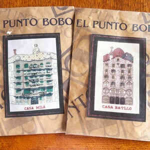 May include: Two cross-stitch kits featuring architectural designs. One kit depicts Casa Milà, the other Casa Batlló. Both kits are packaged in brown paper with the text "EL PUNTO BOBO" printed on the packaging. The Casa Milà kit has green accents, while the Casa Batlló kit has red accents.