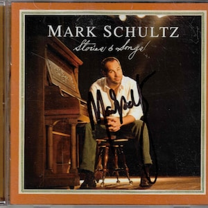 May include: A CD cover featuring a man sitting at a piano. The cover is titled "Mark Schultz Stories & Songs!" and is signed by the artist.